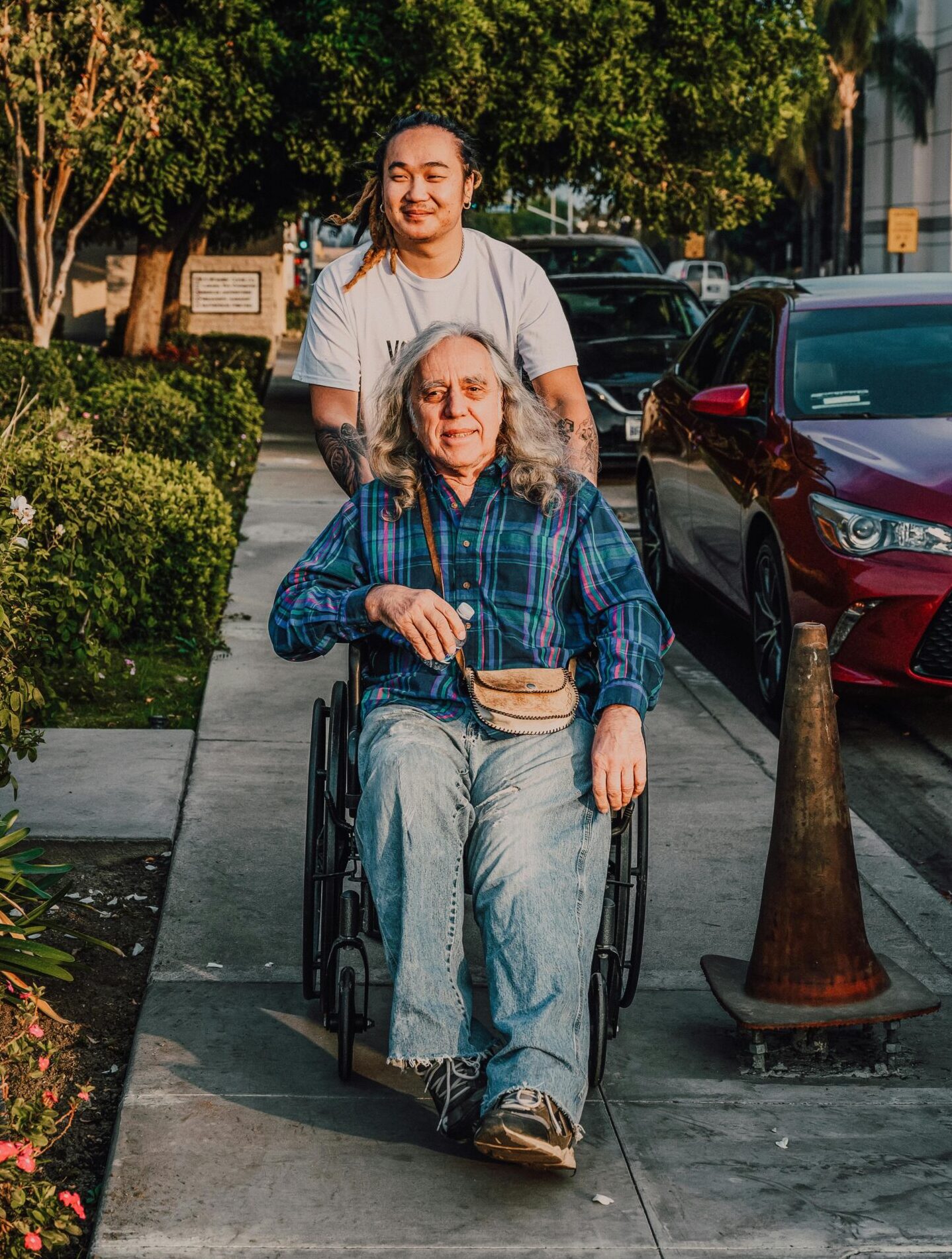 A caring scene of a young man pushing a senior in a wheelchair along a sidewalk.