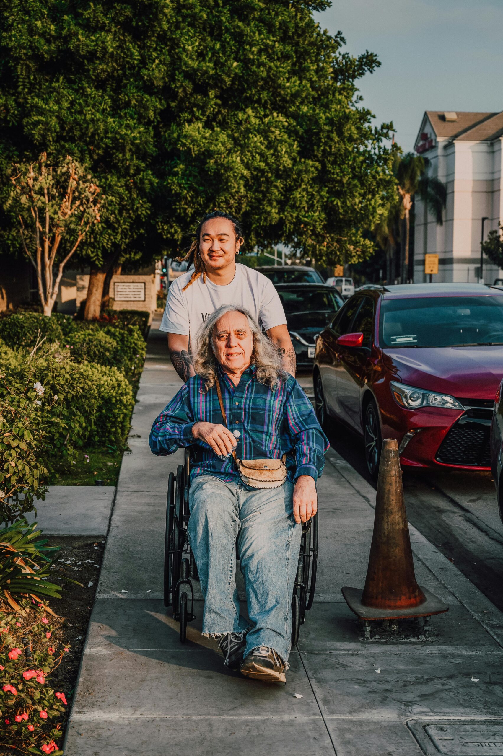 A caring scene of a young man pushing a senior in a wheelchair along a sidewalk.