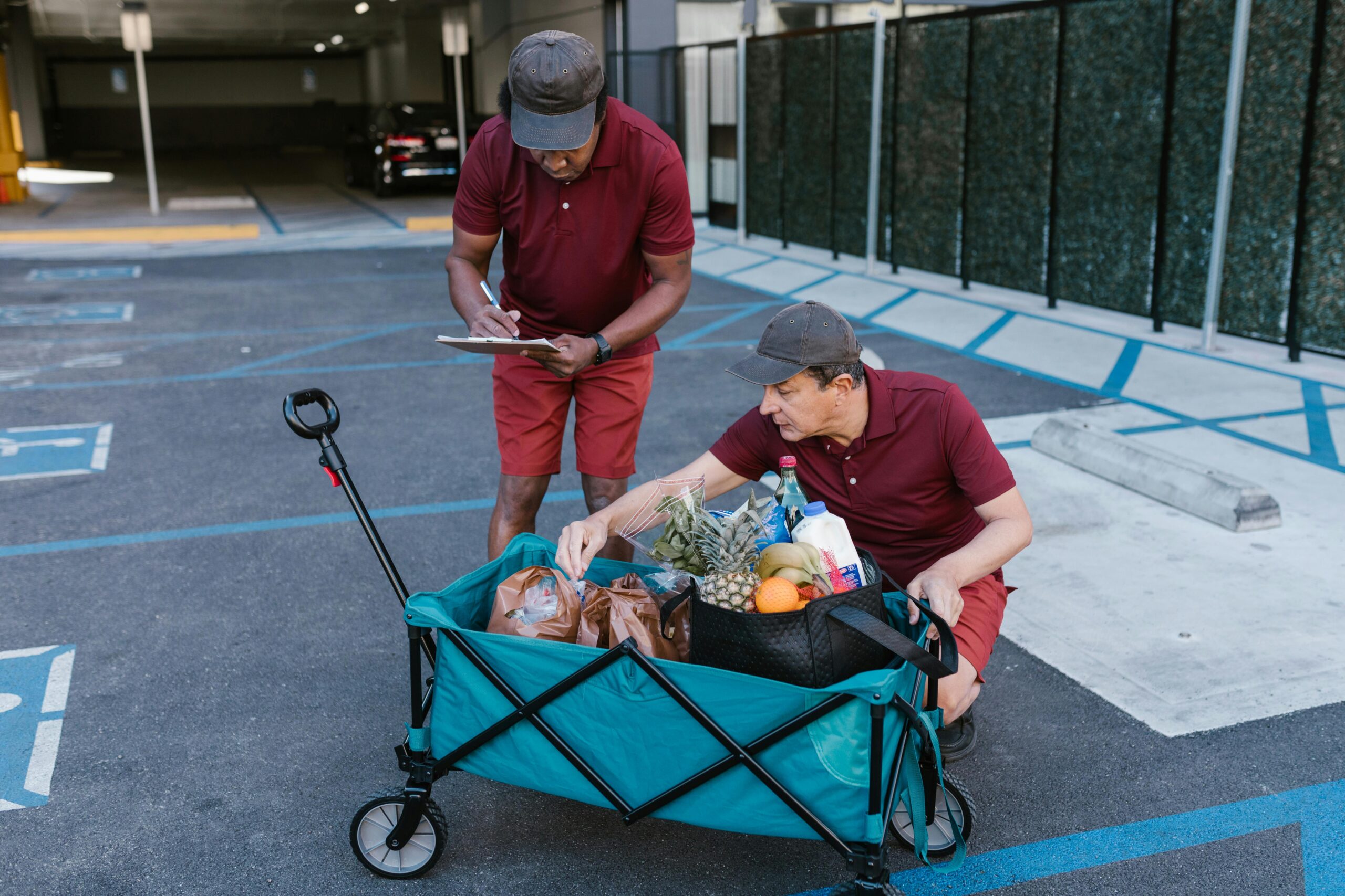 Two men in red uniforms delivering groceries with a push cart in a parking area.